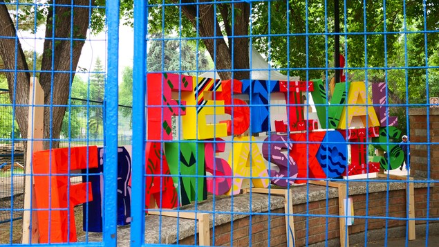 Une installation colorée derrière une clôture bleue, composée de grandes lettres en bois décorées et multicolores formant les mots Festival Fransaskois.