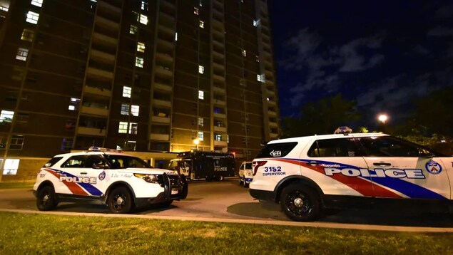 Photo de deux véhicules de police blancs, bleus et rouges stationnés devant un immeuble en briques de plusieurs étages, le soir.