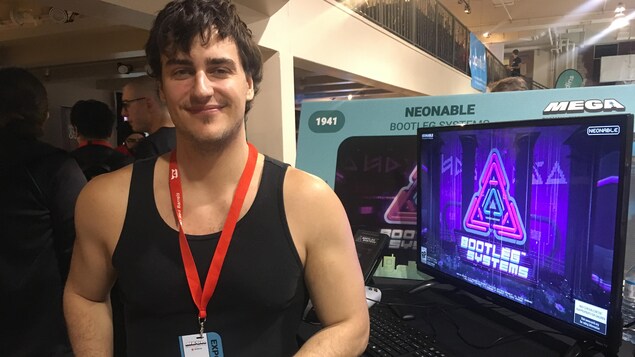 Un homme en camisole noir regarde la caméra en souriant. Il a le bras appuyé sur un bureau sur lequel se trouve un écran sur lequel est affiché le logo d'un jeu vidéo. Un homme en camisole noir regarde la caméra en souriant. Il a le bras appuyé sur un bureau sur lequel se trouve un écran sur lequel est affiché le logo d'un jeu vidéo.