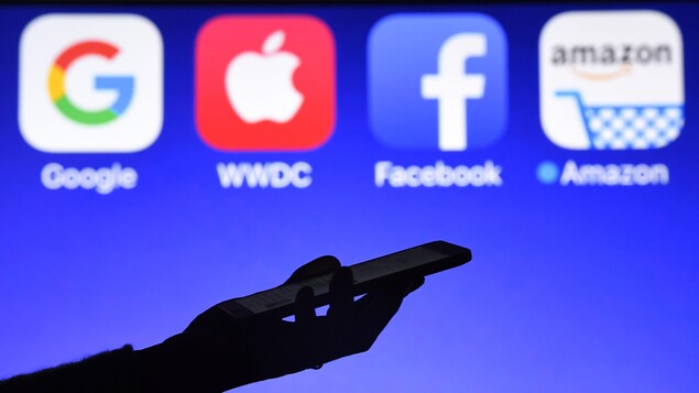 une personne tient un telephone devant les logos de Google, Apple, Facebook et Amazon.