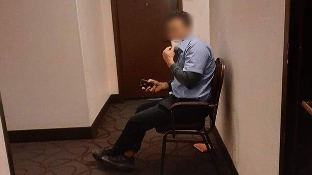 Un homme en chemise avec un masque, assis dans une chaise dans un couloir d'hôtel.