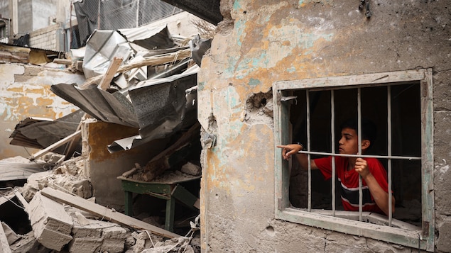 Un garçon derrière une fenêtre avec des barreaux dans une maison qui jouxte des décombres.