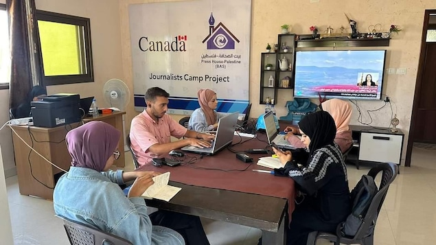 Des femmes et un homme travaillent sur leur ordinateur portable dans un bureau où une affiche «Journalists Camp Project » avec le logo du gouvernement du Canada orne un mur.