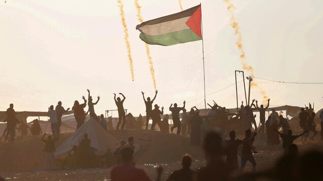 Des Palestiniens lèvent les bras au ciel pendant que des gaz lacrymogènes tombent. Un drapeau palestinien flotte au milieu de la foule.