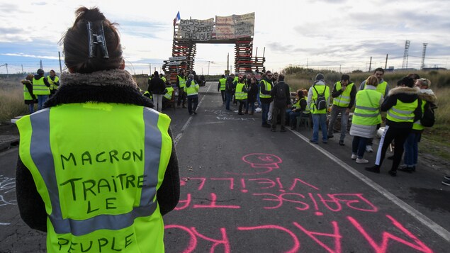 Des « gilets jaunes » bloquent la route. L'un d'entre eux, une femme, est de dos. On peut lire sur son gilet : « Macron, traître. Le peuple a faim. » Des « gilets jaunes » bloquent la route. L'un d'entre eux, une femme, est de dos. On peut lire sur son gilet : « Macron, traître. Le peuple a faim. »