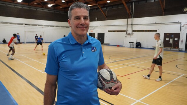 Un homme sourit à la caméra dans un gymnase, avec un ballon de futsal sous la main.