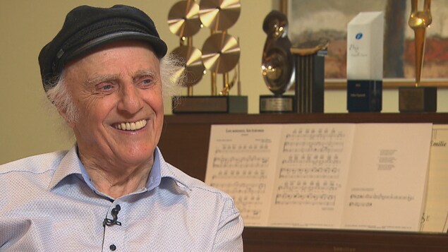 Gilles Vigneault, souriant, assis devant son piano sur lequel reposent ses trophées. Gilles Vigneault, souriant, assis devant son piano sur lequel reposent ses trophées.