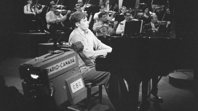 Une caméra filme Glenn Gould qui joue du piano devant un orchestre.