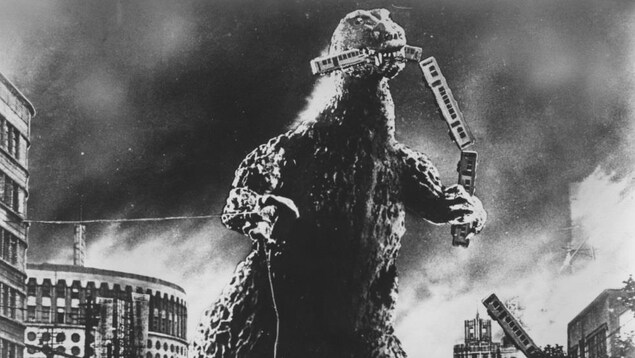 On voit le monstre « Gojira » en train de dévorer un train, dans une ville.