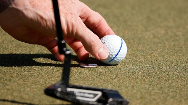 Une balle de golf déposée sur un vert pour une main d'homme avec, à l'avant-plan, la silhouette floue de la tête d'un fer droit