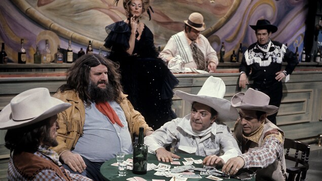Dans un bar, des cowboys (le Grand Antonio, Olivier Guimond et Roger Michael) sont assis autour d'une table et jouent aux cartes pendant que la tenancière (Denise Pelletier), le barman (Rod Tremblay) et un cowboy (Denis Drouin) les observent
au comptoir en arrière-plan. Dans un bar, des cowboys (le Grand Antonio, Olivier Guimond et Roger Michael) sont assis autour d'une table et jouent aux cartes pendant que la tenancière (Denise Pelletier), le barman (Rod Tremblay) et un cowboy (Denis Drouin) les observent
au comptoir en arrière-plan.