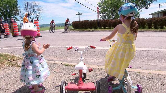 Deux fillettes, l'une à bord de son tricycle, l'autre debout à côté d'elle, regardent les cyclistes passer. Les deux enfants portent une robe et un casque de sécurité. Deux fillettes, l'une à bord de son tricycle, l'autre debout à côté d'elle, regardent les cyclistes passer. Les deux enfants portent une robe et un casque de sécurité.
