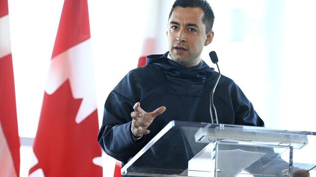 A man stands behind a podium in front of a Canadian flag. 