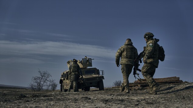 Trois soldats dans un champs austère de dos se dirigent vers leur véhicule armée d'une tourelle. 