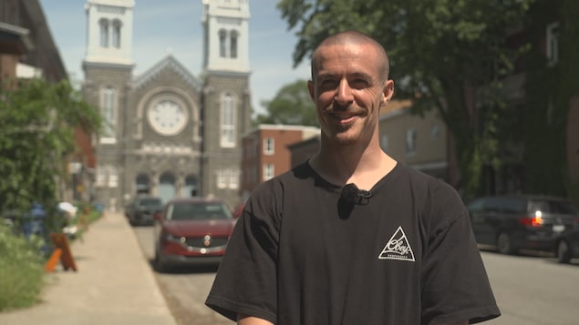 Un homme souriant, au crâne rasé avec une petite barbiche, porte un t-shirt noir avec un logo blanc triangulaire. Il se tient sur un trottoir verdoyant, bordé de maisons et d'une voiture rouge stationnée. À l'arrière-plan, une grande église en pierre grise avec deux clochers domine le paysage urbain sous un ciel clair.