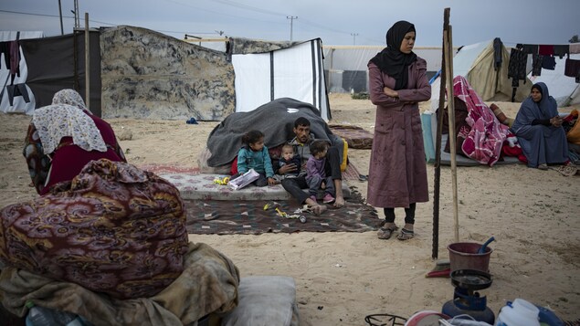 Une femme debout, et à l'arrière, un homme assis sur un matelas posé sur un tapis avec trois enfants, en plein air, dans ce qui ressemble à un camp de réfugiés. 