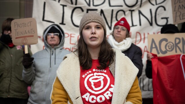 Une femme vêtue d'un t-shirt route décoré du logo d'ACORN en Nouvelle-Écosse, est debout devant un groupe de personnes dans les marches de l'hôtel de ville, pendant une manifestation pour les droits des locataires.