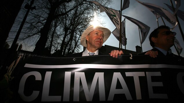 James Hansen, portant un chapeau, derrière une banderole sur laquelle on ne peut que lire le mot climat, en anglais, en lettres majuscules.