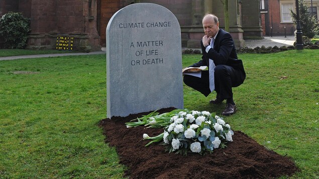 James Hansen est accroupi à côté d'une fausse pierre tombale sur laquelle il est écrit « Changement climatique : une question de vie ou de mort » en anglais. 