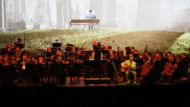 Un orchestre symphonique est sur scène. Derrière lui, la projection d'une image d'un homme sur un banc de parc. 