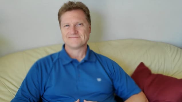 Thomas Hartle assis sur un canapé, les mains croisées et souriant à la caméra.