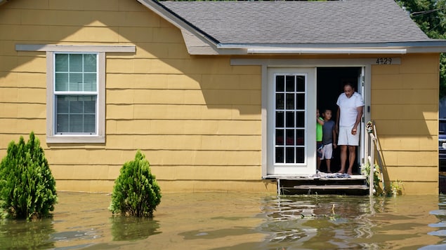 Un homme et ses enfants se tiennent dans le cadre de porte de leur maison jaune, les yeux vers l'eau qui entoure la résidence et monte pratiquement jusqu'à leurs pieds.