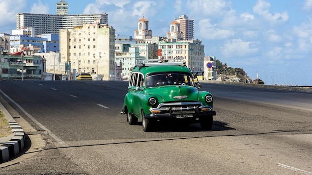 Une vieille voiture roule sur une route déserte. En arrière-plan, une ville avec de vieux bâtiments et des édifices récents.