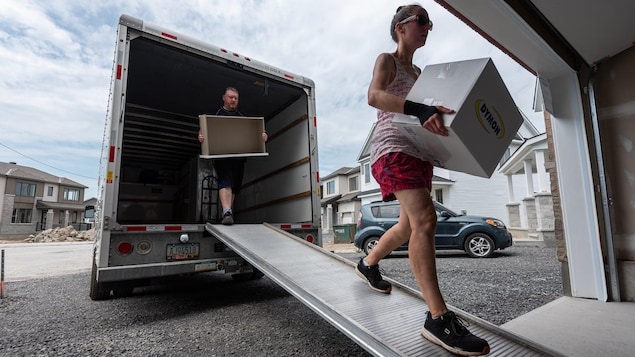 A man and a woman holding a box and a piece of furniture walk down the ramp of a moving truck.