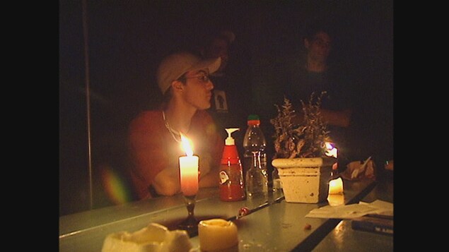 Trois jeunes hommes autour d'une table à l'extérieur, dans la pénombre. Sur la table, des bougies allumées. Trois jeunes hommes autour d'une table à l'extérieur, dans la pénombre. Sur la table, des bougies allumées.
