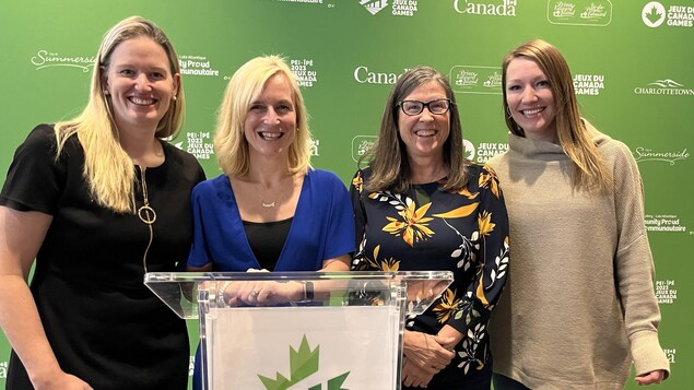 Sami Jo Small, Heather Morrison, Vicki Keith et Heather Moyse posent en souriant pour une photo entre un grand mur vert et un podium décoré du logo des Jeux du Canada 2023.
