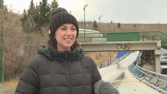 Helen Upperton au bord de la piste de bobsleigh avec les anneaux olympiques en arrière-plan. Helen Upperton au bord de la piste de bobsleigh avec les anneaux olympiques en arrière-plan.