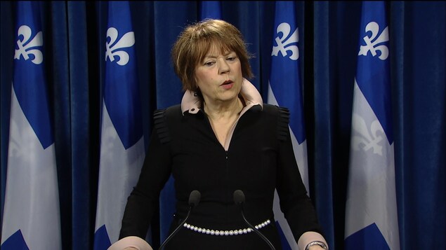 Hélène David devant un micro et des drapeaux du Québec