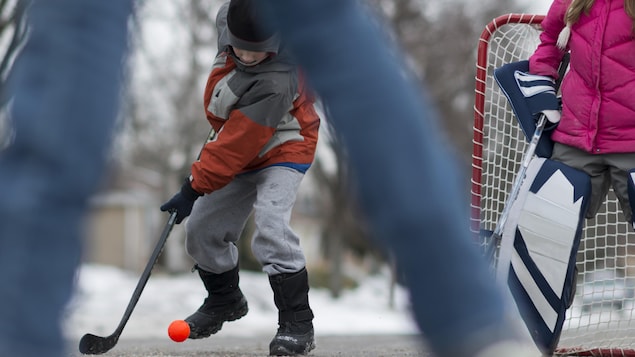 Des enfants jouent au hockey dans la rue en hiver. Un garçon d'âge primaire tente de contrôler une balle rouge avec un bâton de hockey. Une fillette est devant un but posé dans la rue.