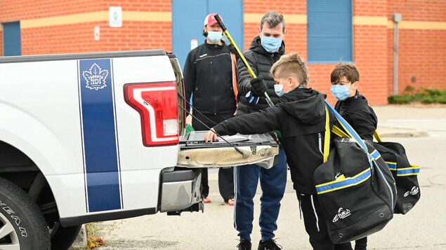 Des enfants et un père déposent du matériel de hockey à l'arrière d'un camion. Ils portent tous un masque.