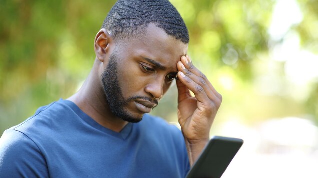 Un homme noir, l’air inquiet, consulte son téléphone intelligent dans un parc.