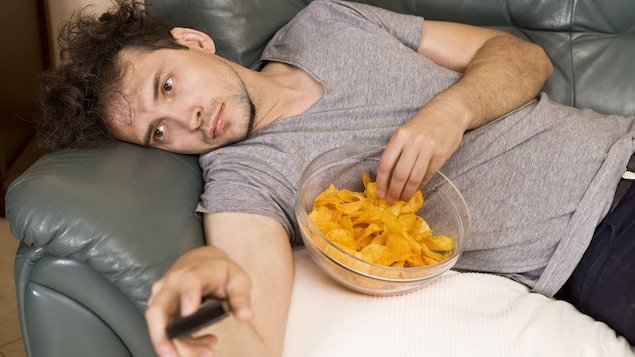 Un homme allongé sur un sofa mange des croustilles en regardant la télévision.