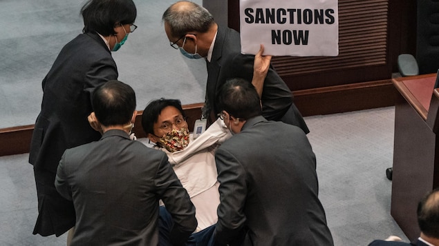 « Des sanctions maintenant », peut-on lire sur une pancarte tenue par un député masqué, qui est tenu par les bras et les jambes par quatre autres personnes.