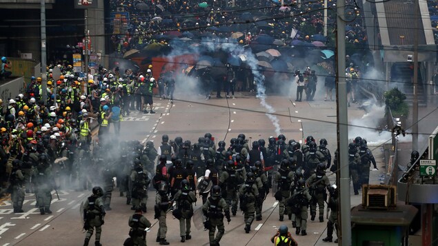 Une foule de manifestants avec des parapluies sont à une vingtaine de mètres de dizaines de policiers en tenue antiémeute qui lancent des gaz lacrymogènes.