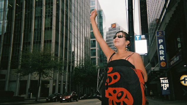 Une femme vêtue d'une robe traditionnelle autochtone noire et rouge lève la main en signe de victoire au centre-ville de Vancouver au bord de la rue avec en arrière plan de grandes tours. Une femme vêtue d'une robe traditionnelle autochtone noire et rouge lève la main en signe de victoire au centre-ville de Vancouver au bord de la rue avec en arrière plan de grandes tours.
