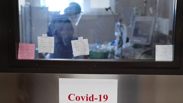 Une personne qui travaille à l'hôpital est vue à travers la fenêtre de l'unité des soins intensifs. Des notes sur de petits papiers et une affiche qui indique « Covid-19 » sont collées sur la porte et la fenêtre de la salle.