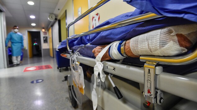 Le bras d'un patient couché sur une civière dans le couloir d'un hôpital. On voit au loin un travailleur médical vêtu d'une combinaison bleue.