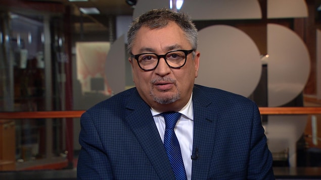 Un homme portant des lunettes et un veston bleu marin regarde la caméra lors d'un entrevue télévisée dans le studio de Radio-Canada.