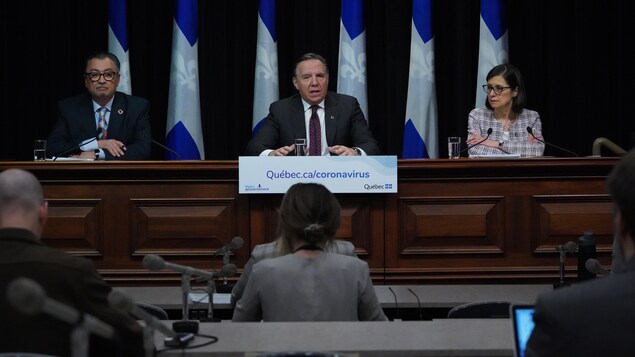 Deux hommes et une femme assis à une table en conférence de presse devant des drapeaux. fleurdelisés.