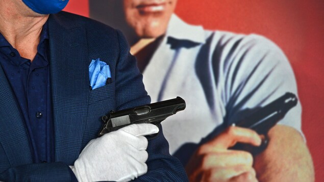 Un homme masqué et ganté tient un pistolet, devant une affiche de Sean Connery dans le rôle de James Bond tenant le même pistolet.