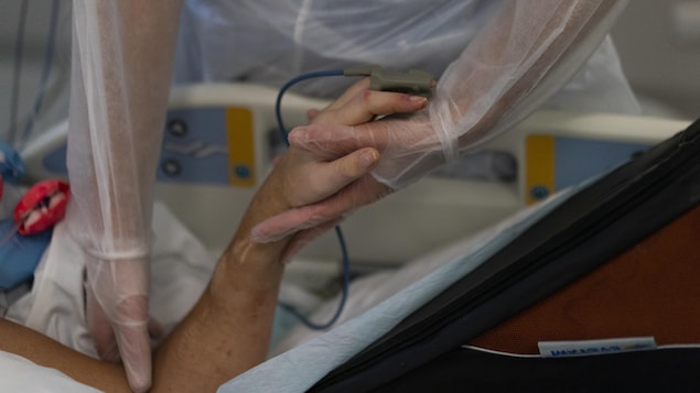 Plan rapproché du bras d'un patient couché dans un lit d'hôpital. Une infirmière lui tient la main.