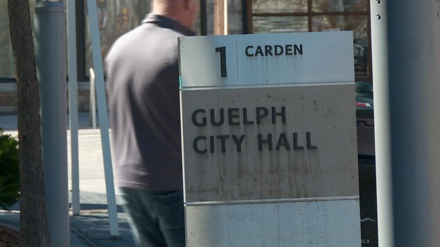On voit une pancarte à l'entrée de l'hôtel de ville sur laquelle est inscrit ''Guelph City Hall". Un homme marche derrière. On voit une pancarte à l'entrée de l'hôtel de ville sur laquelle est inscrit ''Guelph City Hall". Un homme marche derrière.
