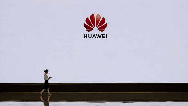 Une femme marche devant un écran blanc sur lequel est projeté le logo de la compagnie Huawei.