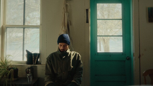 Une image tirée du film, le personnage, au regard triste, assis contre le mur de la cuisine d'une maison de campagne. 