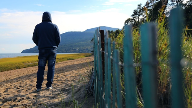 A man wearing a hoodie with his back to the camera looks at the horizon on a beach, by a fence.