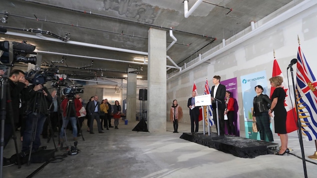Des journalistes et leurs caméras font face à un podium où prennent place des officiels et derrière lequel se trouvent des drapeaux du Canada et de la Colombie-Britannique. Des journalistes et leurs caméras font face à un podium où prennent place des officiels et derrière lequel se trouvent des drapeaux du Canada et de la Colombie-Britannique.